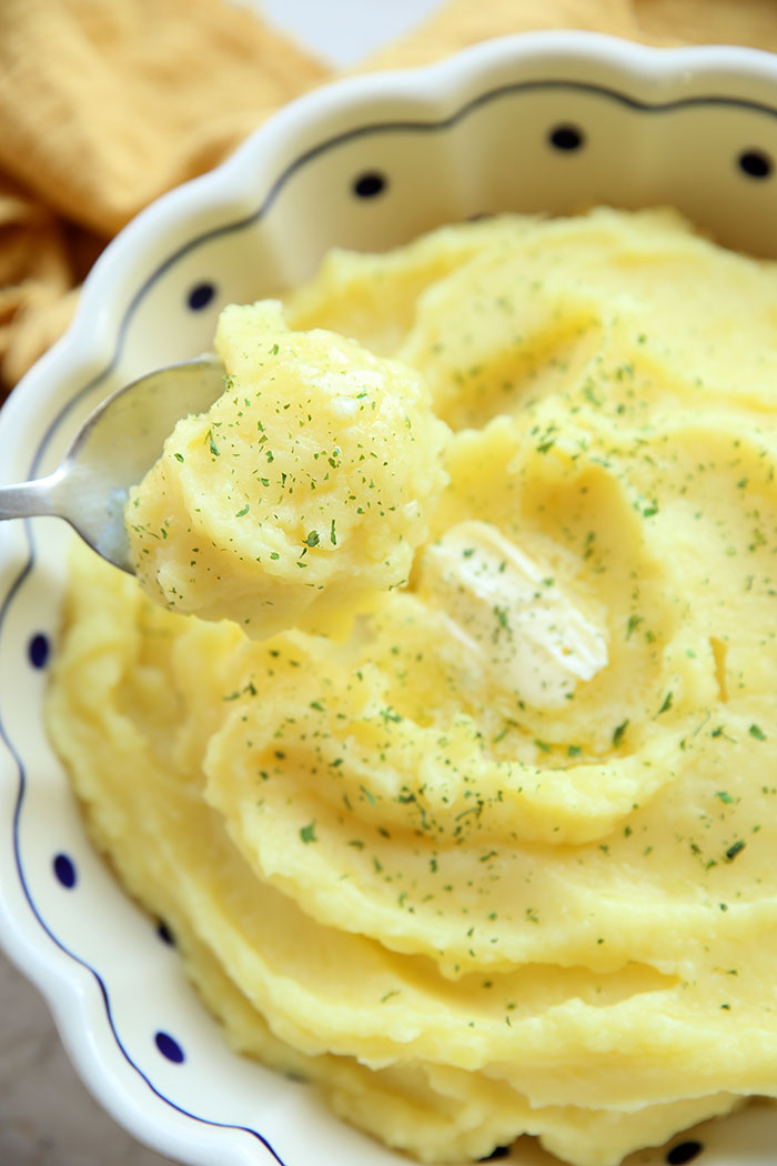 Mashed Potatoes in a white scalloped bowl with a blue trim and polkadots. A spoon is held above the dish with a scoop of mashed potatoes in it. Above the bowl is a cloth yellow napkin laid loosely down.