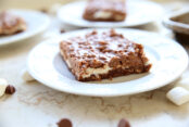 A piece of marshmallow crunch brownie sitting on a white dessert plate with small marshmallows and milk chocolate chips scattered around the plate. In the background, there's two small white plates with a brownie on top and a corner of a metal cookie sheet.