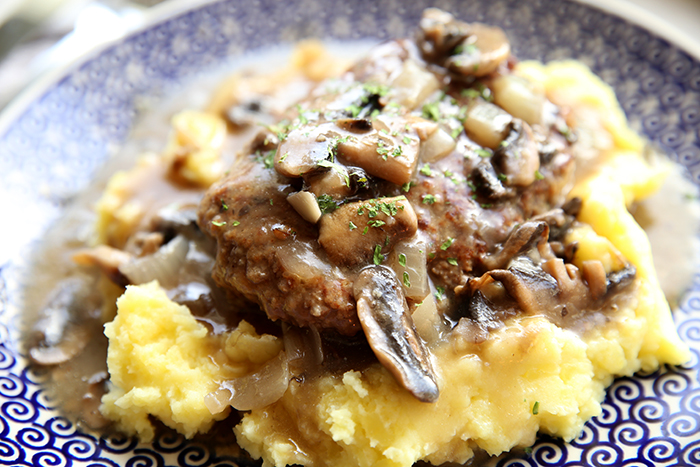 A blue plate with a serving of salisbury steak with mushroom and onion gravy on top of buttermilk mashed potatoes. 