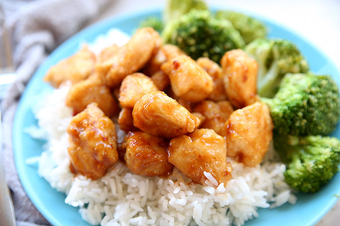 Orange chicken sitting on a serving of white rice next to a serving of broccoli all on top of a blue plate. In the left background is a grey kitchen towel.
