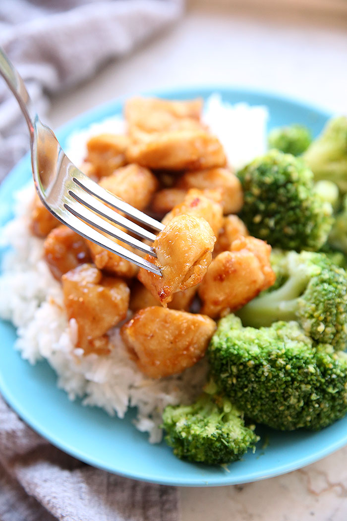 A light blue plate with white rice topped with orange chicken and broccoli. Above the chicken a fork is starting to lift a piece of chicken up. In the background, there's a gray and white striped kitchen towel.