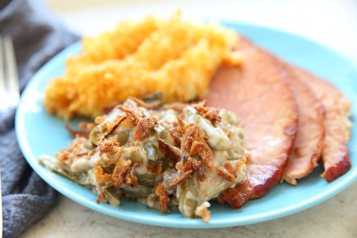 A serving of Classic Green Bean Casserole and funeral potatoes with three slices of ham all on a light blue plate. On the left of the plate, there's a blue kitchen towel with a partial picture of a fork head.