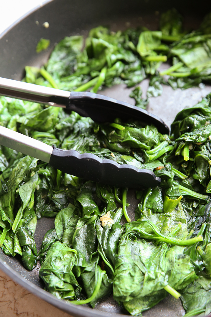 A vertical photo showing a partial picture of a large frying pan of sautéed spinach. A pair of rubber tipped tongs is getting ready to take a bunch out from the left side of the pan.