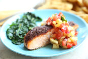 A light blue plate with a serving of oven baked blackened salmon topped with pineapple salsa with a serving of garlic sautéed spinach. Blurred in the top background is a fork and a yellow cloth kitchen towel.