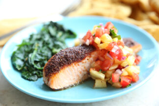 A light blue plate with a serving of oven baked blackened salmon topped with pineapple salsa with a serving of garlic sautéed spinach. Blurred in the top background is a fork and a yellow cloth kitchen towel.