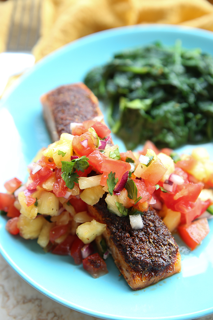 A blue dinner plate with a piece of blackened salmon topped with pineapple salsa with a serving of sautéed spinach. In the top background is a yellow dish towel and the head of a fork.