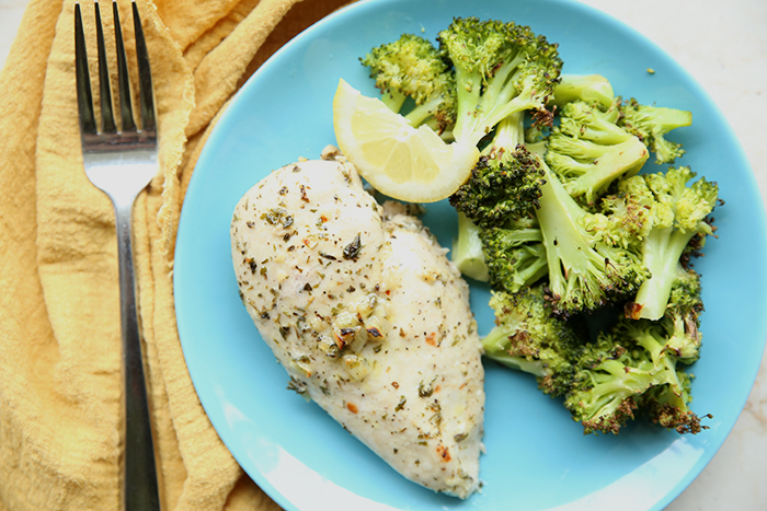 A serving of steamed broccoli, a lemon wedge and a lemon herb chicken breast all sitting on a blue plate. A fork is on the left of the plate sitting on a golden cloth kitchen towel.