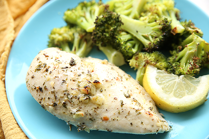 A light blue dinner plate with a piece of lemon herb chicken, a serving of broccoli and a lemon wedge. On the left of the plate is a golden cloth kitchen towel.