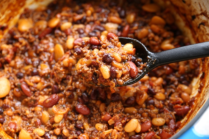 A large cooking pot filled with baked beans recipe. On the right side a black serving spoon is in the pot with a scoop of beans and held above the rest of the beans.