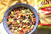 A white and dark blue patterned bowl with blue polka dots full of black bean avocado salsa. On the right of the bowl is a bag of tortilla chips. Above the blue on the left is a loosely laid golden cloth napkin.