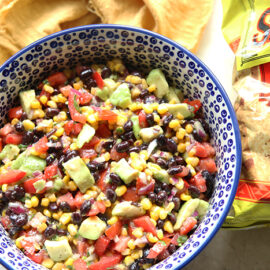 A white and dark blue patterned bowl with blue polka dots full of black bean avocado salsa. On the right of the bowl is a bag of tortilla chips. Above the blue on the left is a loosely laid golden cloth napkin.