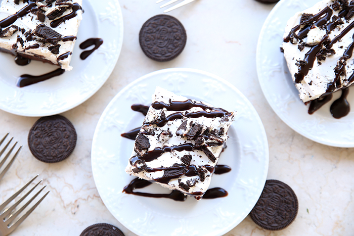 Three servings of Brownie Oreo Ice Cream Cake with Hershey's chocolate sauce drizzled on top. Two servings are in the upper corners with one in the middle. Oreos are scattered around the plates with two fork heads in the left bottom corner.