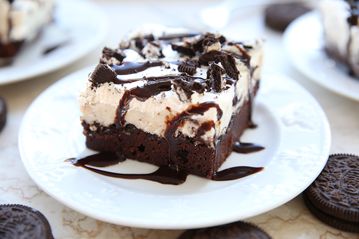 A square piece of brown ice cream cake on top of a dessert plate drizzled with Hershey's syrup. Scattered around the plate are Oreos with two servings of cake faded in the upper corners.
