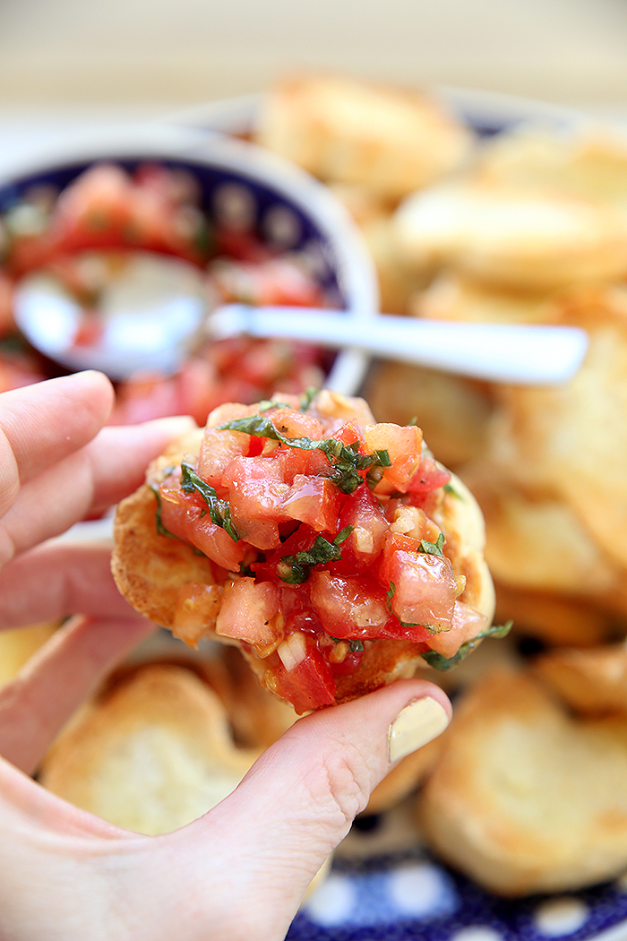 A hand is holding a piece of ciabatta bread topped with bruschetta. Faded in the background is a plate with slices of toasted bread and a small bowl with bruschetta and a spoon in it.