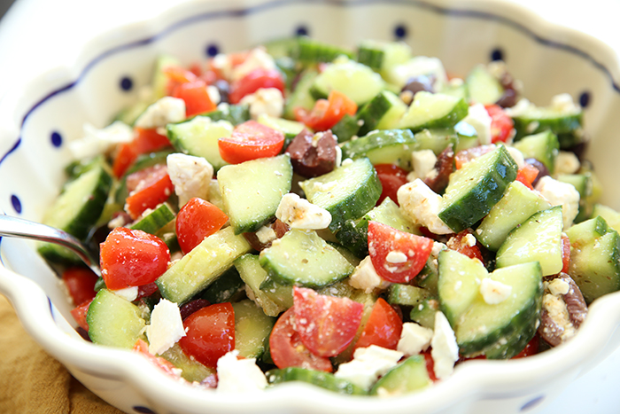 A white bowl with a scalloped edge with dark blue dots filled with Easy Greek Salad recipe. A metal spoon is inserted into the salad on the left side.