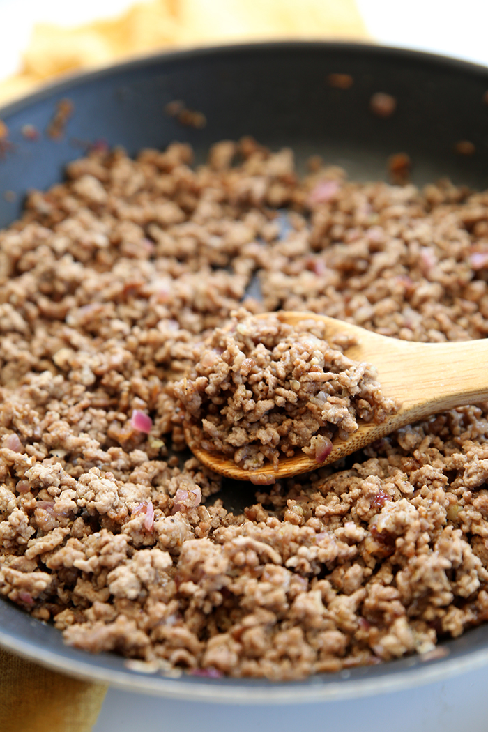 A large frying pan of Greek ground beef with a wooden serving spoon lying in the middle and on the edge of the pan with a scoop of ground beef.