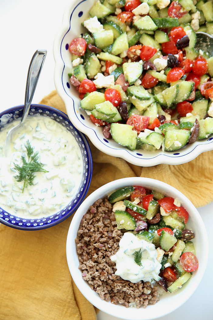 A large scalloped white Polish pottery bowl with blue dots filled with Greek Salad. Below the large bowl is a small white bowl filled with ground beef, Greek Salad, and a dollop of tzatziki sauce. Next to the large bowl is a small dark blue bowl with white dots filled with tzatziki sauce with a sprig of dill in the middle and a small spoon inserted into the sauce. Underneath all the bowls is a golden cloth hand towel.