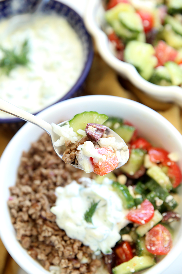A spoon with a scoop of Greek Bowl held over a small white bowl filled with Greek salad, Greek ground beef, topped off with a scoop of tzatziki sauce. In the upper faded background is a dark blue bowl with tzatziki sauce with a sprig of dill in the middle and a partial picture of a scalloped edged bowl of cucumber, tomato, and feta salad.