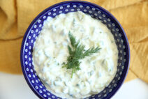 A dark blue bowl with white polka dots filled with Greek Tzatziki Sauce with a sprig of dill on top of the sauce in the middle. Underneath the bowl is a yellow cloth kitchen towel.