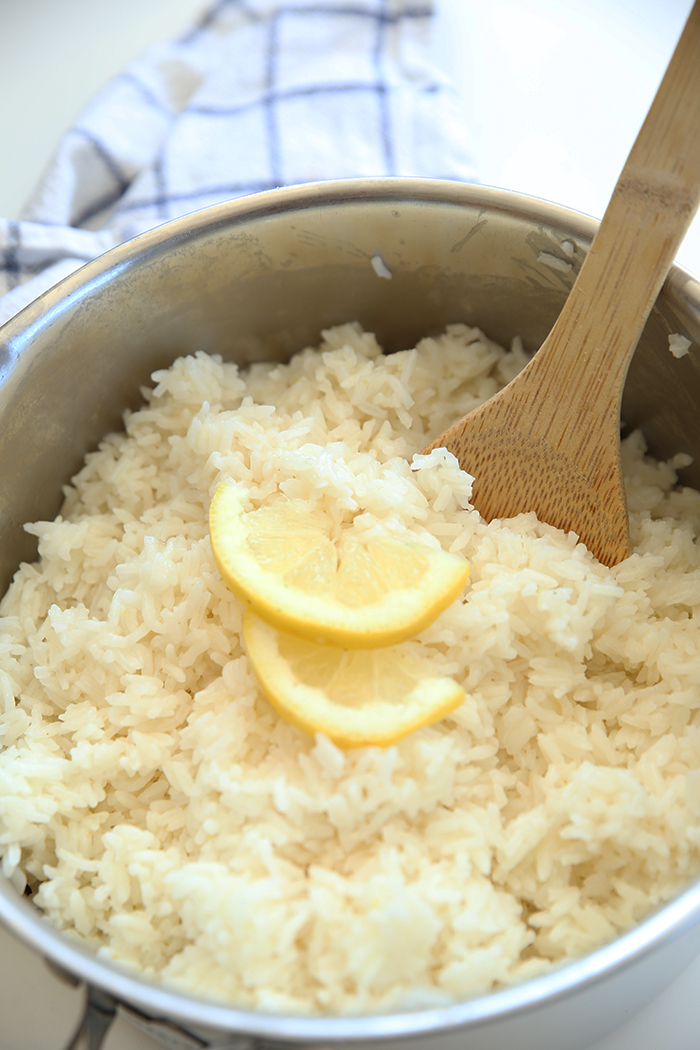 A large metal pot filled with lemon flavored rice with a wooden serving spoon in the rice upright along with two slices of lemon. A white kitchen towel with a blue checkered pattern is at the top in the background.