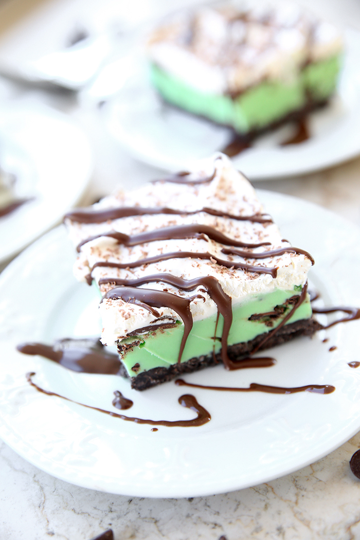 A slice of mint chocolate chip ice cream cake on a small white plate topped with grated chocolate and drizzled with chocolate syrup. In the faded background is another piece of cake on a small white plate and a partial picture of a small white plate.