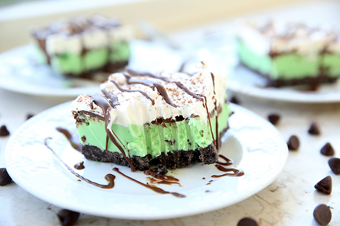 A slice of mint chip ice cream cake on top of a white dessert plate with grated chocolate and chocolate syrup drizzled on top and the plate. A bite of cake is taken out of the corner of the cake. In the faded background are two more pieces of cake on small white plates. Chocolate chips are scattered around the plates.