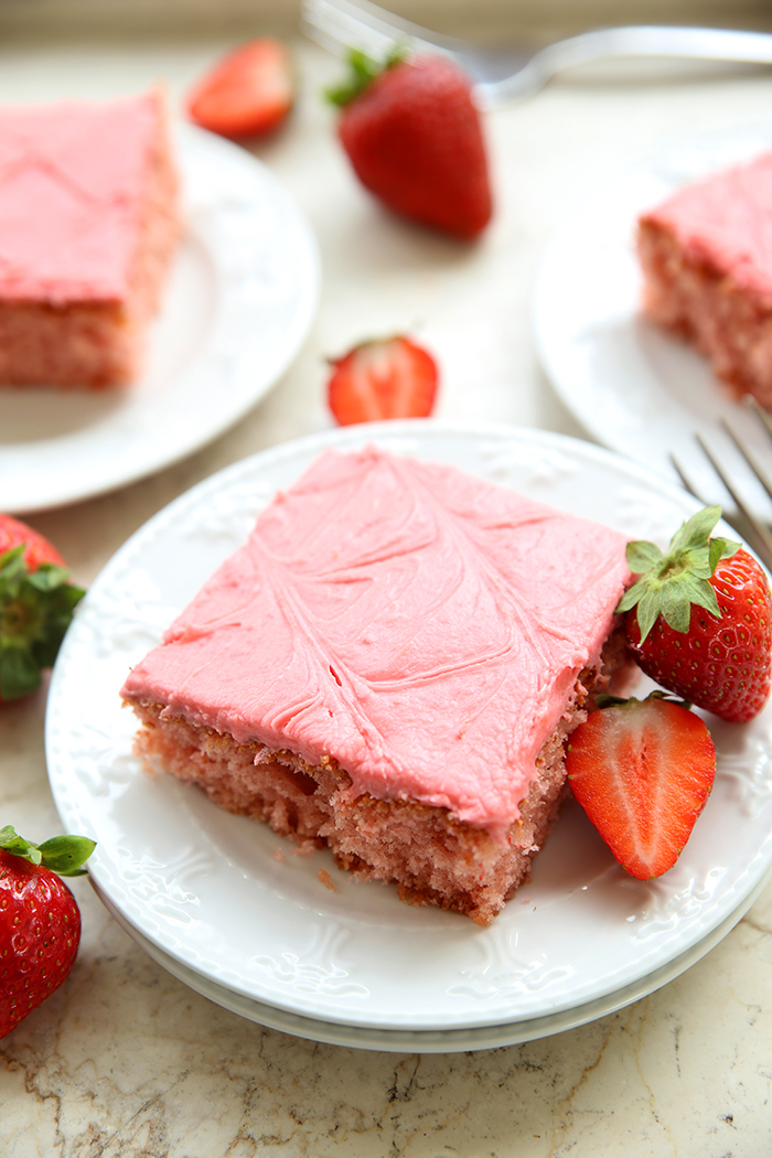 large piece of pink strawberry cake on a white plate with a sliced straberrry on the side and a few more plates and pieces visible in the background