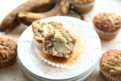 A toddler cream cheese muffin cut in half with butter smeared on it sitting on a stack of small white plates. Several muffins surround the plates along with two ripe bananas.