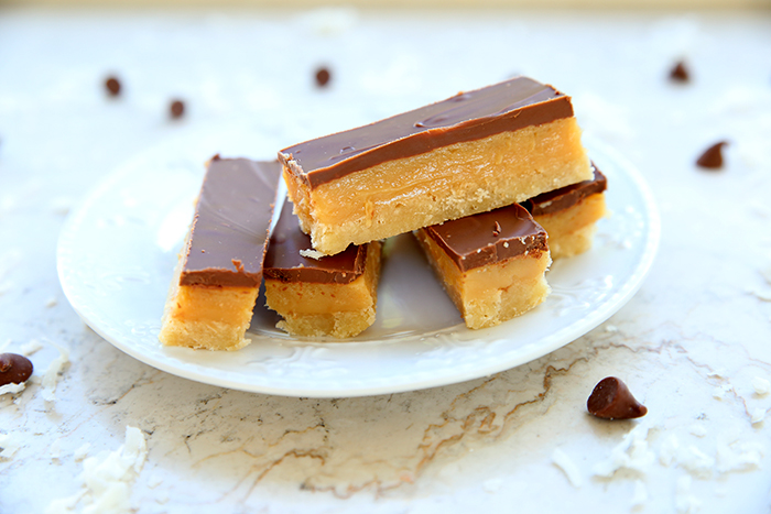 Five pieces of caramel slice on a dessert plate with coconut and chocolate chips scattered around the plate.