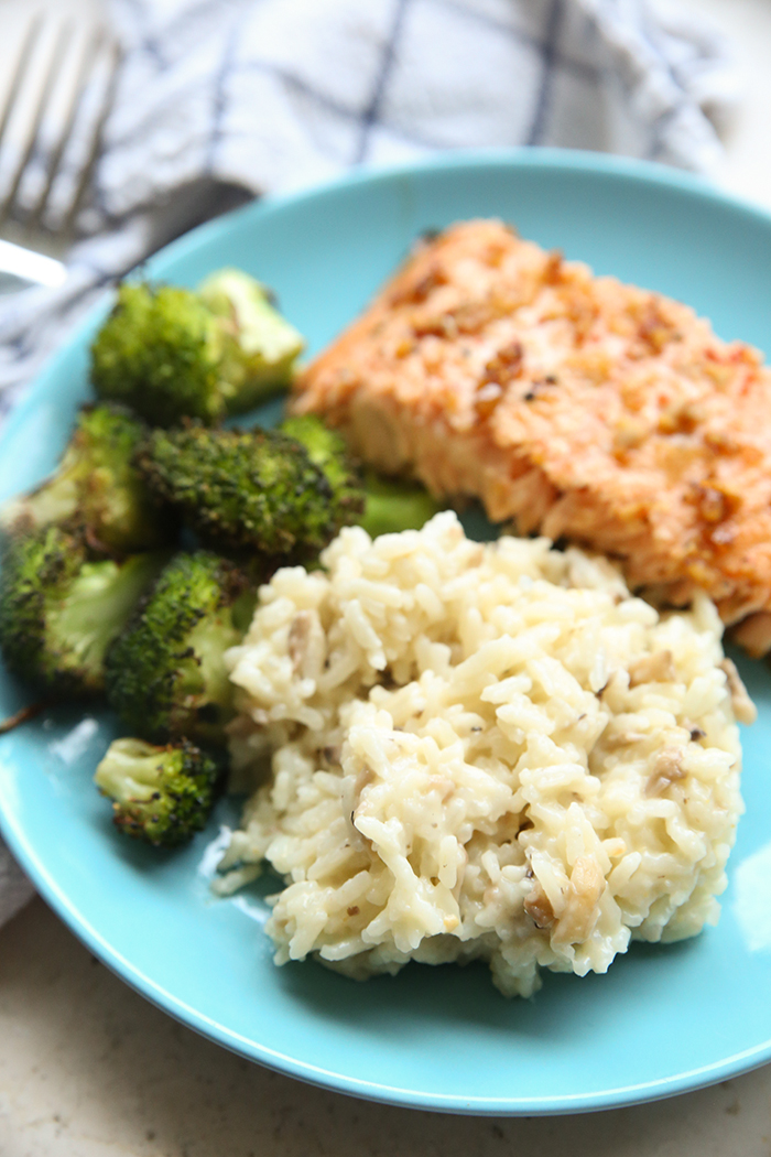 Instant Pot Mushroom Risotto on a blue plate with broccoli and salmon. In the upper background is a white and blue striped kitchen towel with a fork head in the left upper corner.