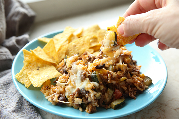 A serving of taco skillet on a plate with corn chips. A hand is holding a corn chip scooping up some zucchini and beef.