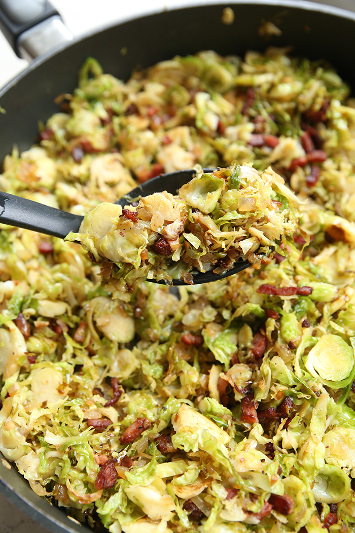 Partial picture of a large skillet of shredded Brussel sprouts with bacon. A serving spoon is held above the pan with a scoop of brussel sprouts.