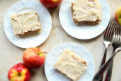 Three small plates with square pieces of apple sheet cake with brown butter frosting. Two whole apples are in the right bottom corner, and three forks are laid on top of each other on the left side.