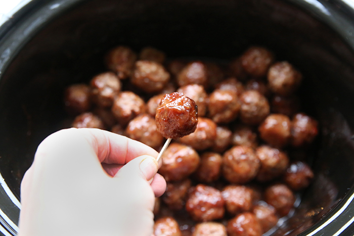 A slow cooker of meatballs with a hand held above holding a meatball with a toothpick.