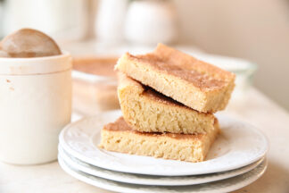 Three slices of Snickerdoodle Bars stacked on top of three small white plates. To the left of the plates is a cinnamon-sugar shaker.