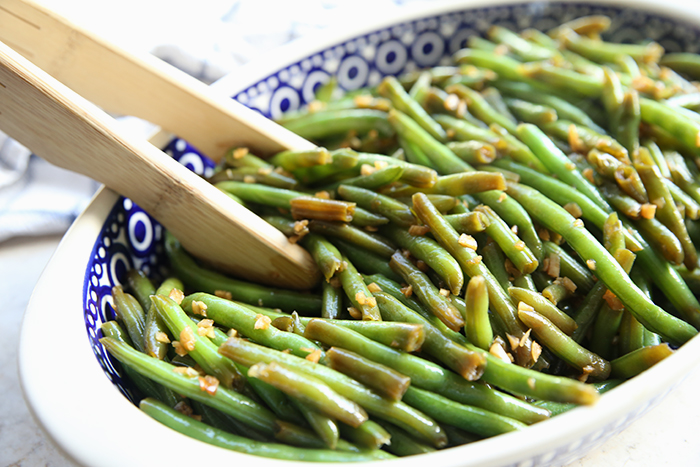 Asian style green beans in a white rimmed peacock patterned Polish pottery dish. A pair of wooden tongs are inserted into the dish on the left side.