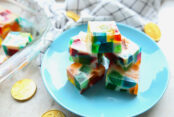 Broken Glass Jello Squares stacked on a small blue plate. Surround the plate are Euro coins. In the upper background is the corner of a glass dish with jello squares and a kitchen towel.
