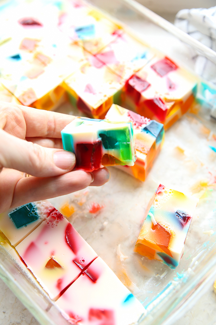 9x13 pan of rainbow jello, there is a hand holding a square with a bite out of it to show how the multiple colors are in each bite.