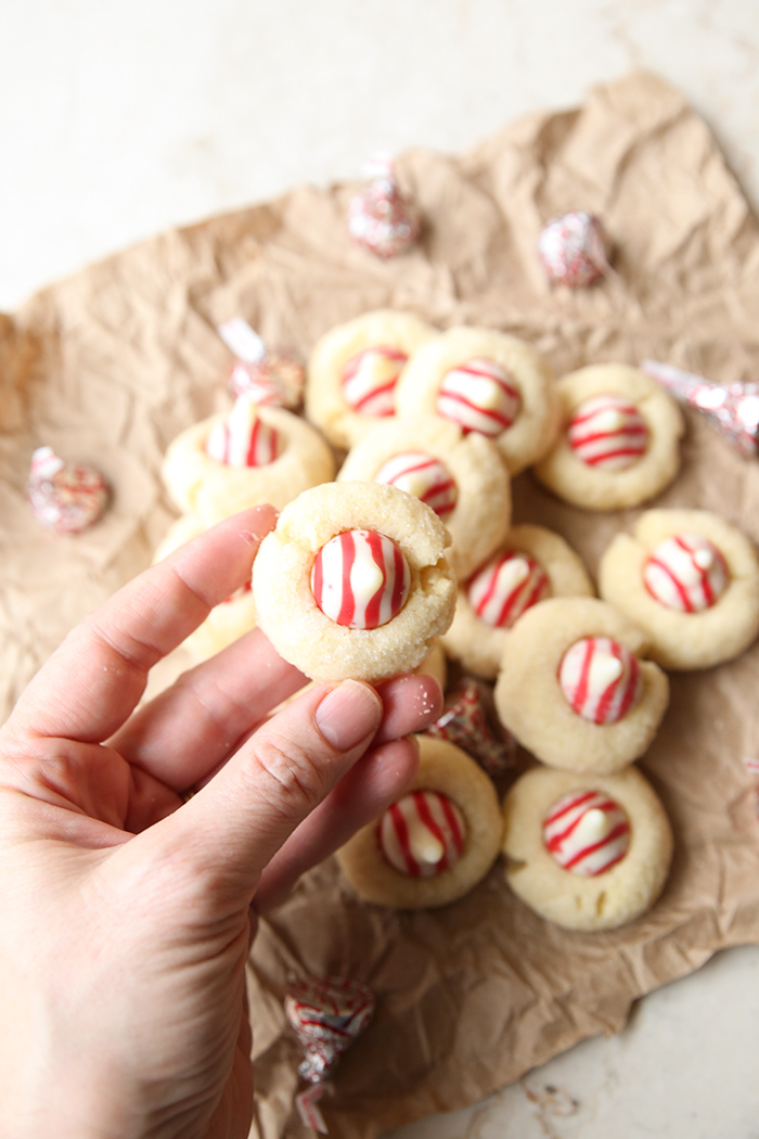 A stack of shortbread peppermint cookies surrounded by Peppermint Hershey Kisses all sitting on a brown crumpled paper bag. A hand held above is holding a peppermint kiss cookie.