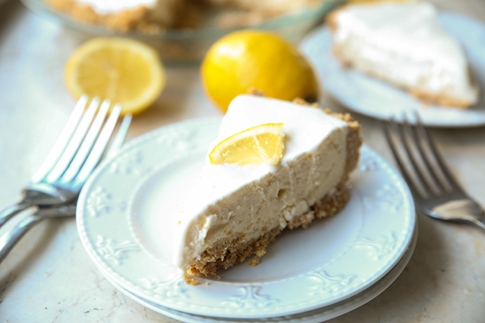 Two small white plates stacked on each other with a slice of three ingredient lemonade ice cream pie with a lemon wedge on top. On the right and left of the plate are forks with half a lemon, a whole lemon, and a piece of pie on a white plate all in the background.