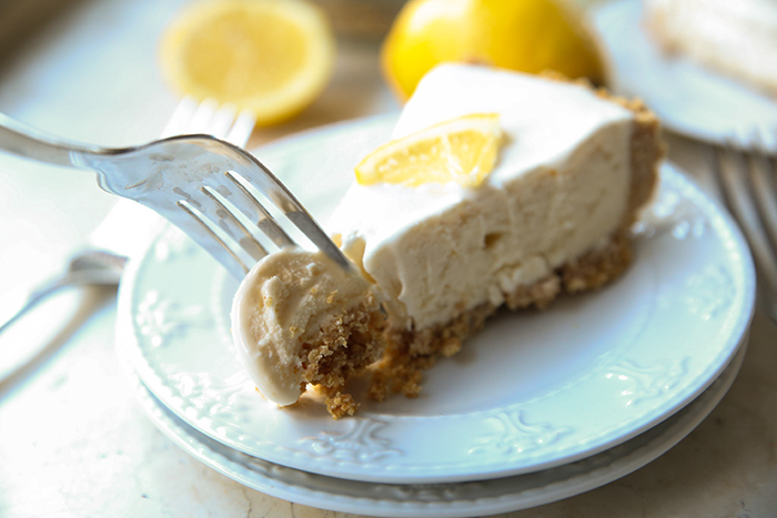 A piece of lemonade ice cream pie with a fork inserted into the tip on two small white plates. In the background are a whole lemon, half a lemon and two forks.