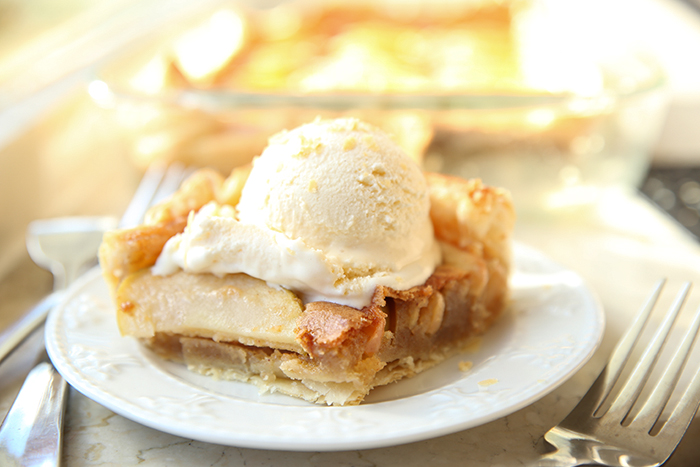 A piece of apple dessert topped with a scoop of vanilla ice cream with forks on both sides of the plate. Faded in the background is a 9x13 dish of Brown Butter Apple Dessert.