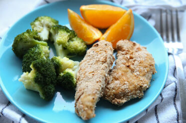A blue plate with two chicken fingers, orange slices and a serving of broccoli. A fork is on the right of the plate sitting on a white and blue striped kitchen towel.