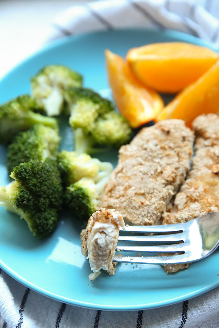 A blue dinner plate with broccoli, orange slices, and two baked chicken fingers. A fork is held above the plate with a piece of chicken on it. A white and blue striped tea towel is under the plate.