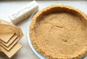 A graham cracker pie crust with an opened package of graham crackers and a stick of butter to the left of it.