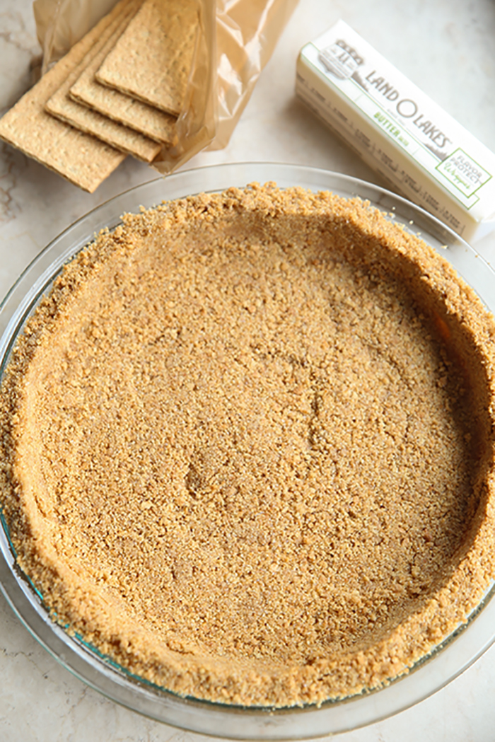A graham cracker pie crust with a package of opened graham crackers and a stick of butter above it.