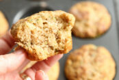 banana zucchini muffin, bikini muffin. Picture of someone holding one they have taken a bite out of it. you can see the muffin tin with more muffins in the background.