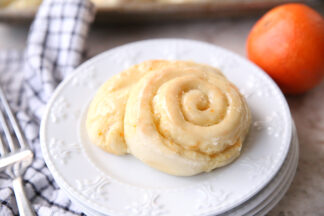 An orange roll sitting on a stack of four small white plates. On the left of the plates is a fork sitting on a white and blacked checked kitchen towel. In the upper right hand corner is a whole orange.