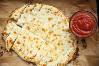 Cheesy garlic breadsticks sitting on parchment paper with a small bowl of pizza sauce on the right side.