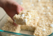 A glass 9x13 dish of cut Rice Krispie Treats with one piece held above by a hand.